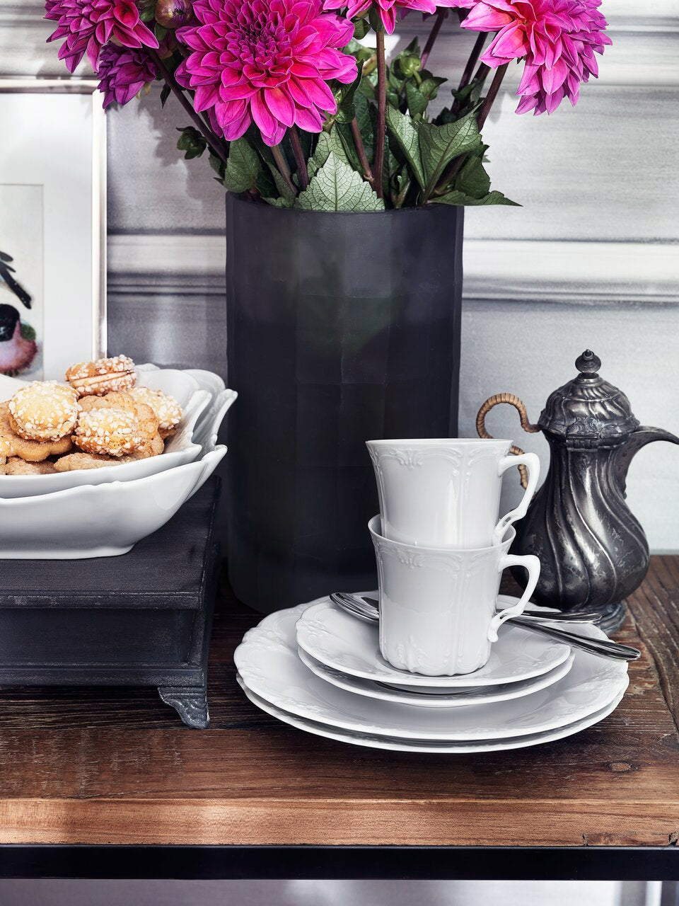 White ceramic cups and saucers on a wooden table with a vase of pink flowers and cookies in the background.