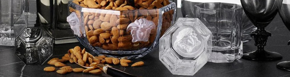 A clear glass bowl filled with almonds sits on a dark countertop, surrounded by matching clear glass containers and a wine glass in a dimly lit setting.