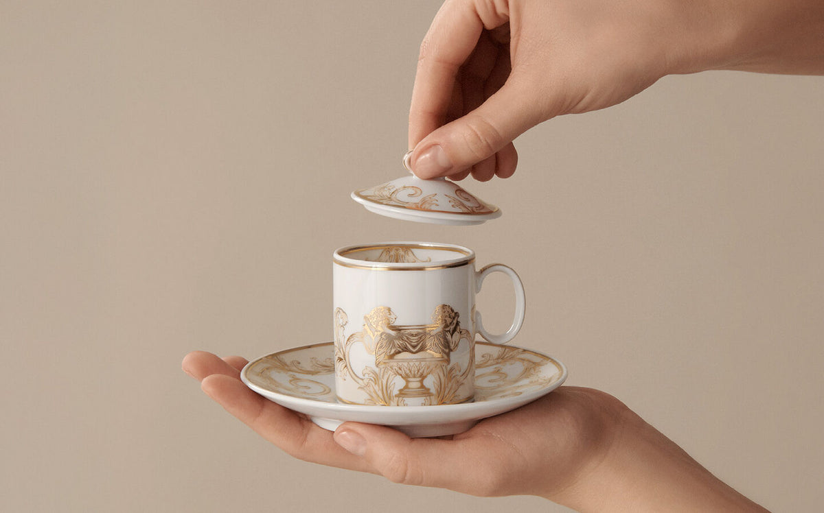 A hand holds a white ceramic teacup and saucer with gold decorative artwork, while another hand lifts the matching lid. The background is a solid, warm beige tone.